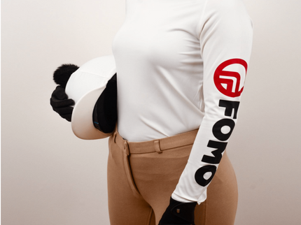 Person wearing a white long-sleeve shirt with a logo, brown riding pants, and black gloves on a plain background.
