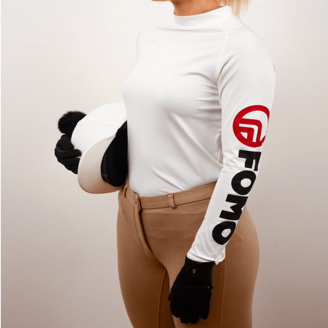 Person wearing a white long-sleeve shirt with a logo, brown riding pants, and black gloves on a plain background.