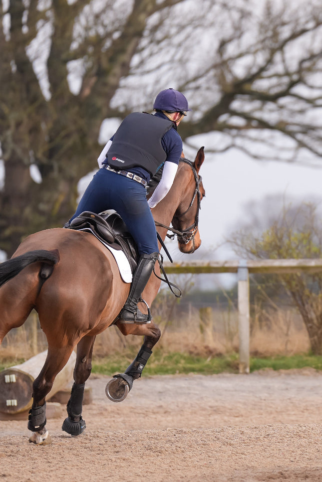 Person riding a horse in an outdoor equestrian setting with trees and a fence in the background.