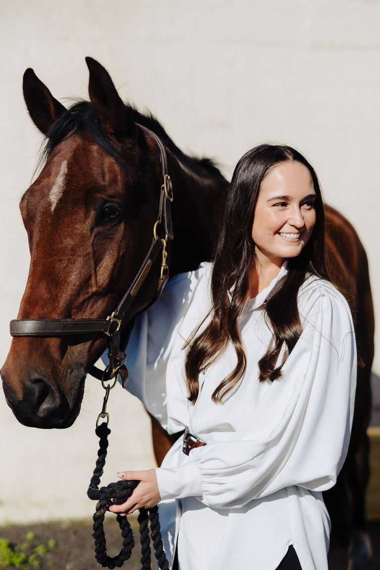 Carmen in a white outfit standing next to a brown horse against a plain background