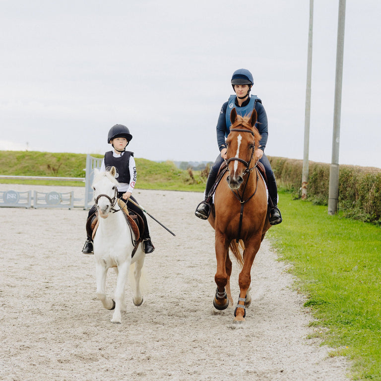 Two people riding horses on a gravel path with grass and a cloudy sky in the background.