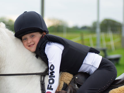 Child riding a white horse wearing a helmet and a vest with 'FOMO' branding.