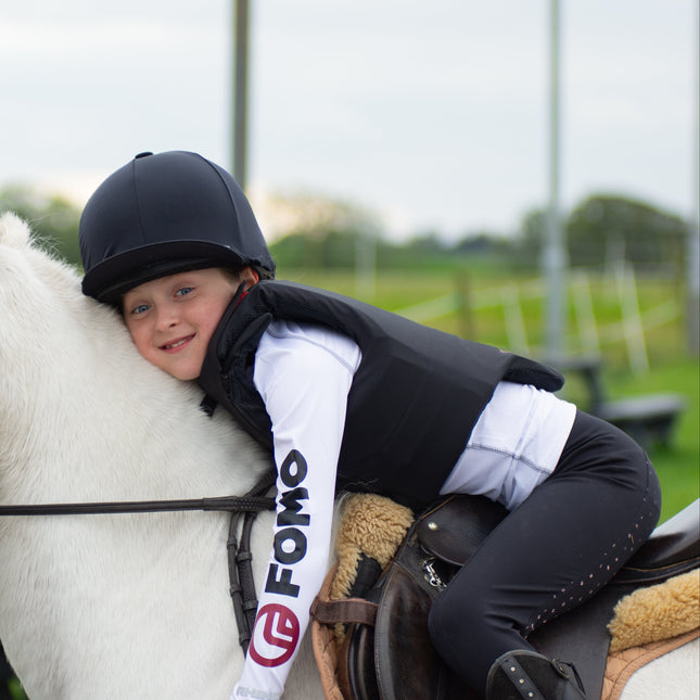 Child riding a white horse wearing a helmet and a vest with 'FOMO' branding.