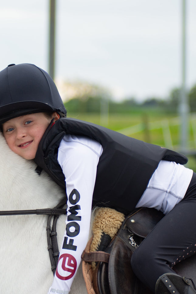 Child riding a white horse wearing a helmet and a vest with 'FOMO' branding.