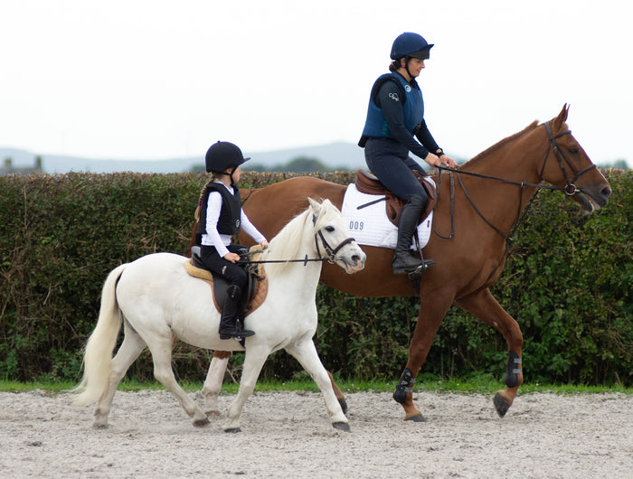 Two people riding horses on a dirt path with a hedge in the background