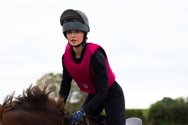 Filitered Person riding a horse wearing a pink vest and helmet on a clear day.